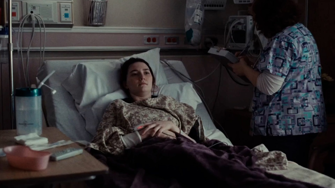 A woman lies in a hospital bed with monitoring equipment nearby. A nurse stands beside her, taking notes.