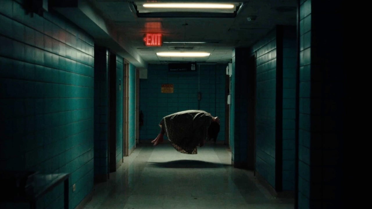 A dimly lit hospital hallway with teal walls and a glowing exit sign. A person in a dress is floating horizontally in the middle of the corridor.