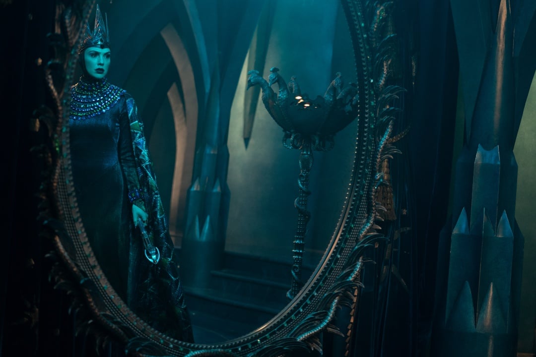 A woman in an elaborate costume and crown stands in an ornate, dimly lit room, reflected in a large decorative mirror.