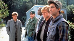 A group of five men in casual clothing stand outdoors near a vehicle, focusing on something in the distance during daylight.