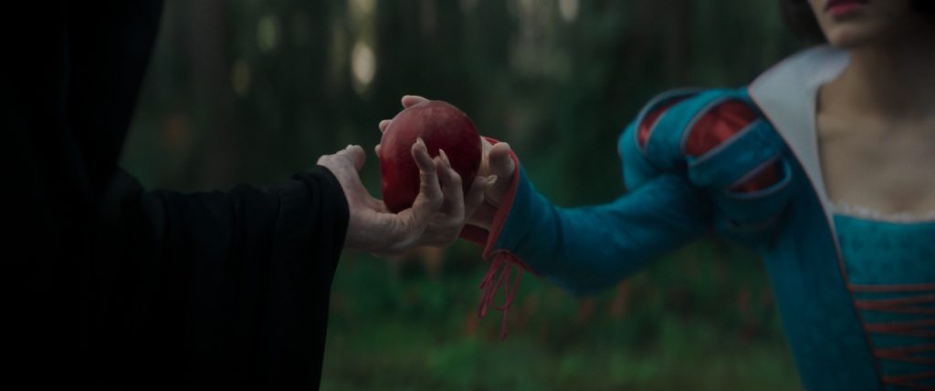 A person in a hooded cloak offers a red apple to another person wearing a blue and red costume in a forest setting.