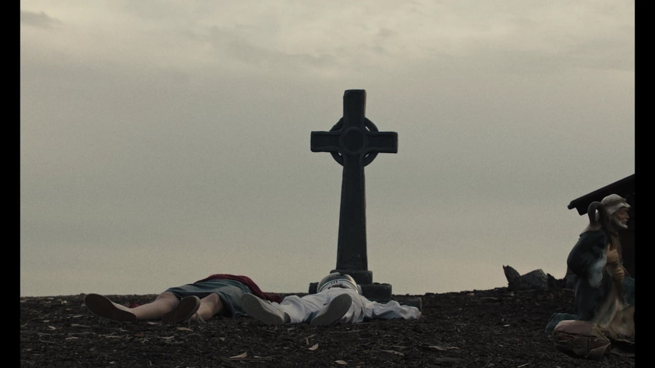 Two people lie on the ground beside a large cross-shaped gravestone. A statue of a shepherd holding a staff is positioned on the right. The sky is overcast.