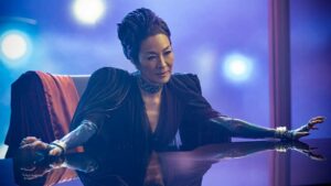 A person wearing a dark, elegant outfit with metallic gloves is seated, leaning forward on a reflective table, set against a blurred background with bright lights.