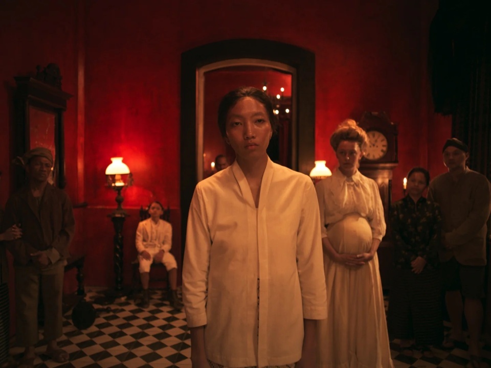 A person in a white shirt stands at the forefront in a dimly lit red room with five people in the background.