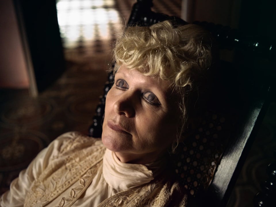An older person with curly blonde hair and a neutral expression sits in a dark, patterned chair wearing a high-collared, light-colored garment.