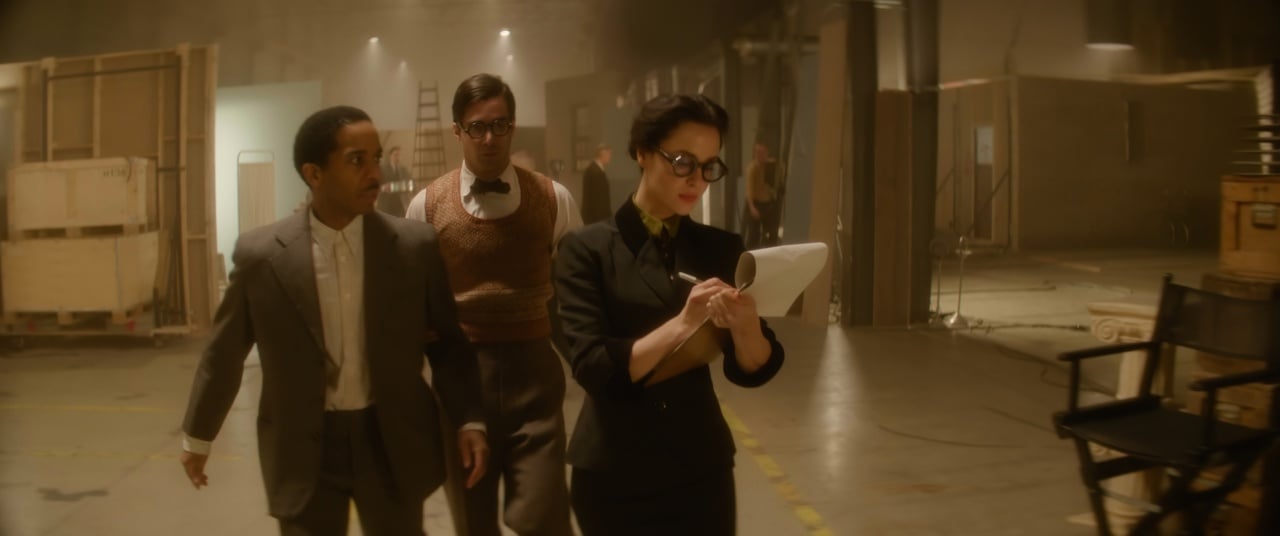 Three people in 1950s-style attire walk together in a large, dimly lit studio. One woman is writing on a clipboard, while two men walk beside her, engaged in conversation.