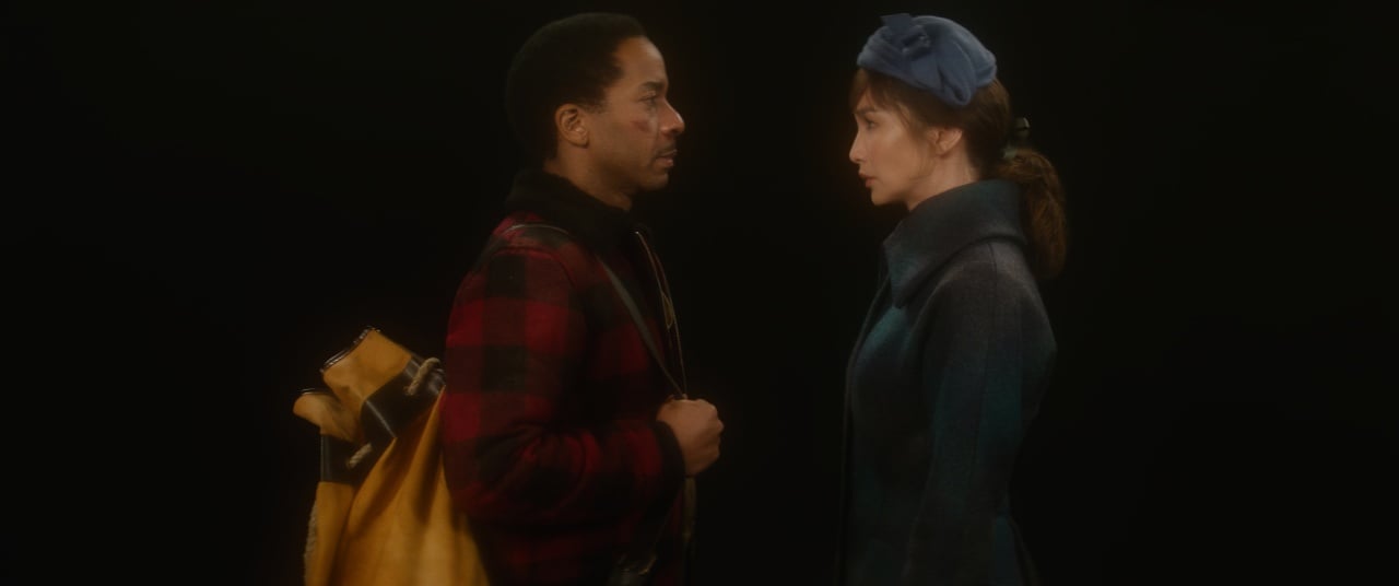 A man and a woman face each other against a dark background. The man wears a red and black checkered jacket and carries a yellow bag. The woman wears a blue coat and hat.