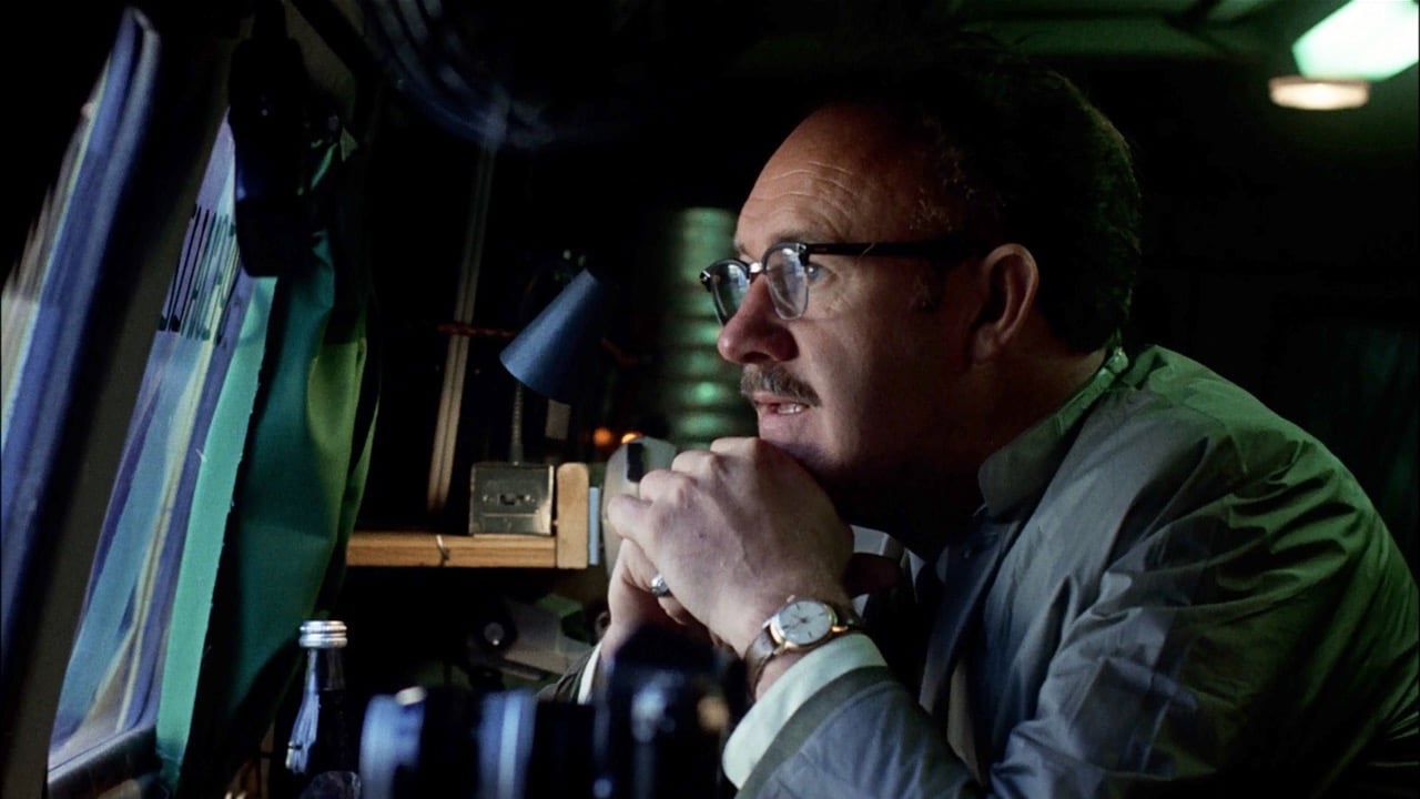 A person wearing glasses and a watch, seated by a window, looks outside while clasping their hands near their mouth.