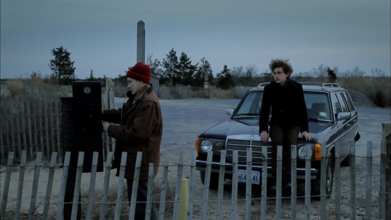Two people are near a parked car by a beach. One person, wearing a red hat, interacts with a black device on a post, while the other sits on the car hood, gazing into the distance.