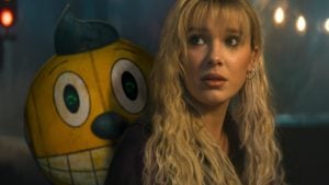 A person with long blonde hair looks into the distance while a large cartoonish yellow character stands behind them in a dimly lit setting.