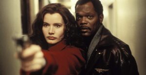 A woman in a red coat points a gun, standing in front of a man in a leather jacket, in a narrow hallway.