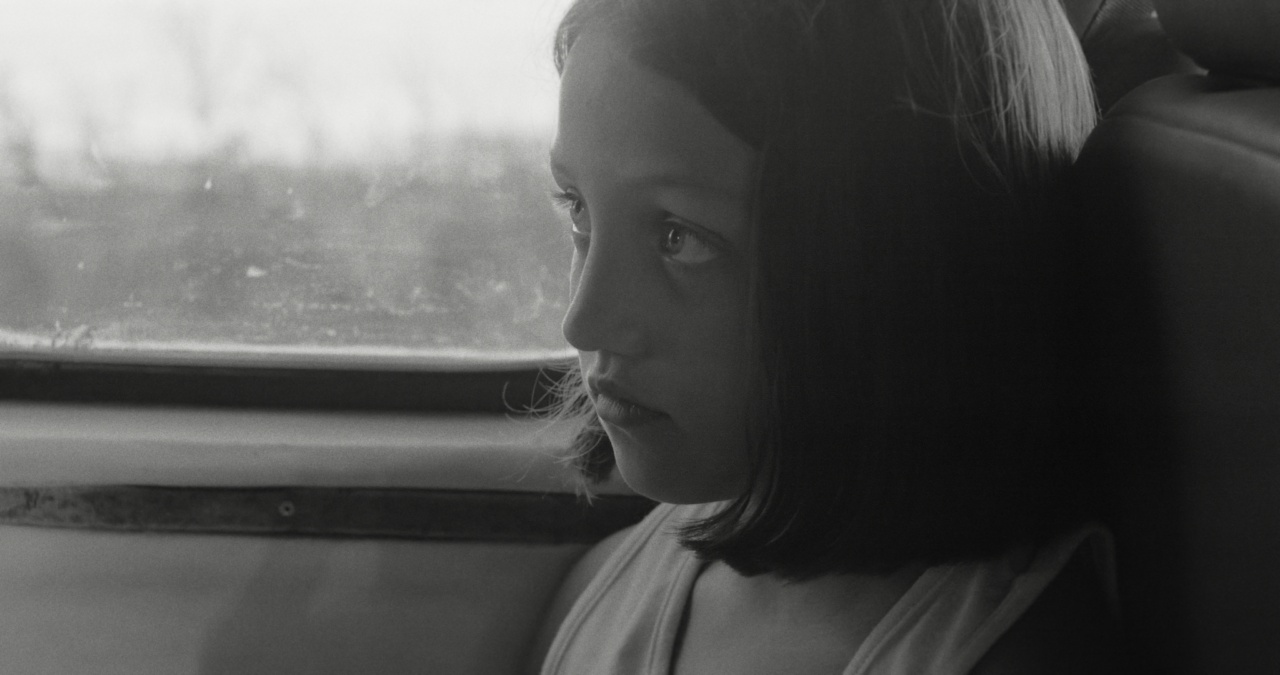 Black and white photo of a young girl with shoulder-length hair sitting by a window, looking outside with a thoughtful expression.