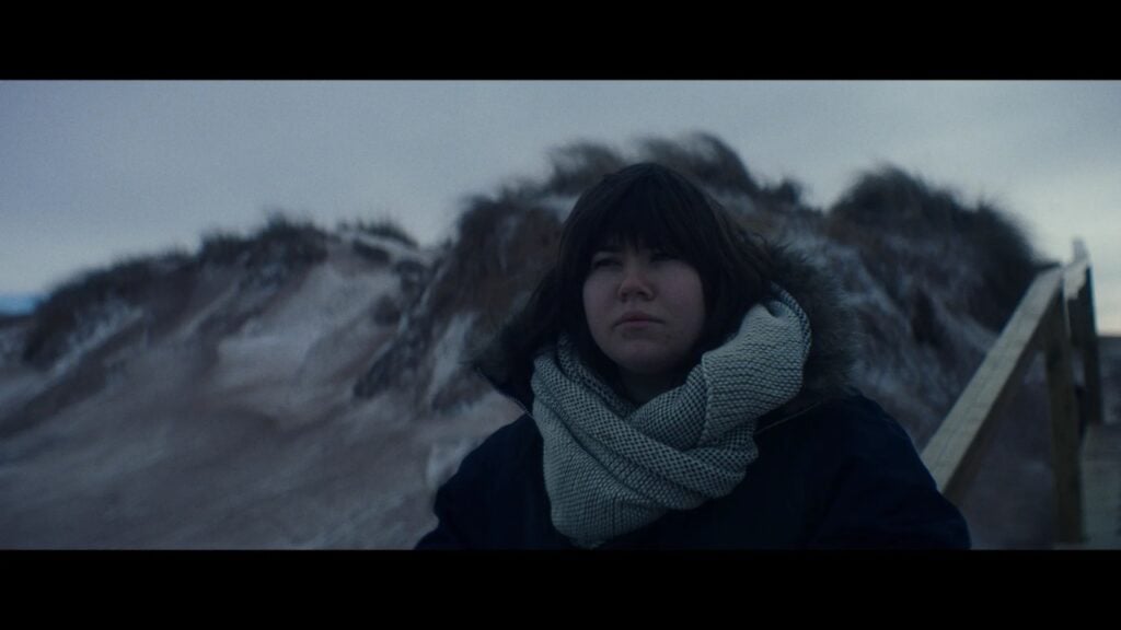 A person with dark hair and a large scarf is sitting outdoors on a wooden pathway, with grass-covered sand dunes in the background under an overcast sky.
