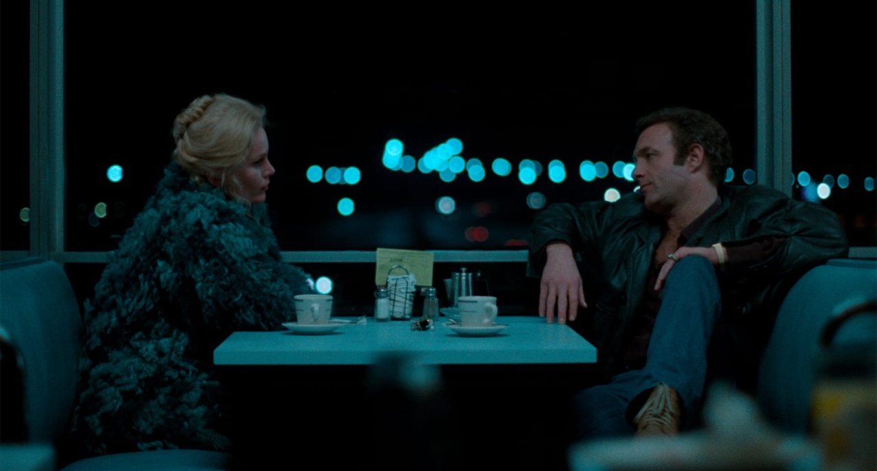 A man and a woman sit across from each other in a dimly lit diner booth, engaged in conversation. Cups of coffee and small items are on the table.