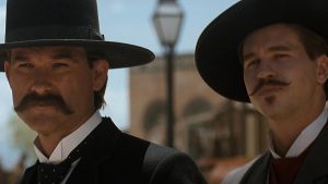 Two men in period attire with wide-brimmed hats and mustaches stand outdoors, with a building and lamp post in the background.
