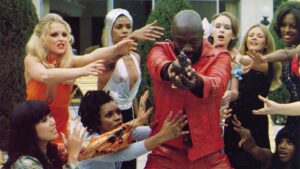 Shout! Studios To Release Debut Volume Of Blaxploitation Classics Box Set On 4K UHD Blu-Ray This May A person in a red outfit points a gun at the camera, surrounded by a group of women reaching towards him with varied expressions.