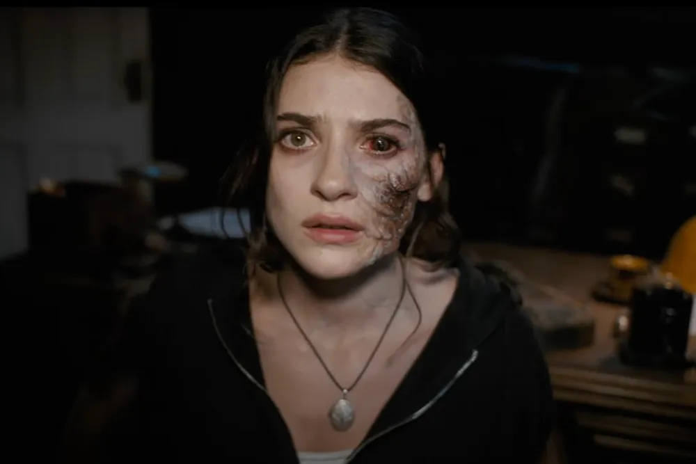 A woman with long dark hair has a serious expression and a scarred face. She is wearing a necklace and a black hoodie in a dimly lit room.