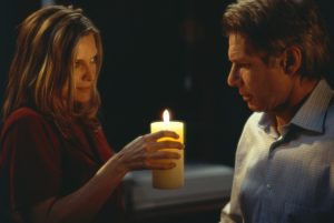 A woman holds a lit candle, looking at a man facing her. Both are indoors, with dim lighting creating a solemn ambiance.