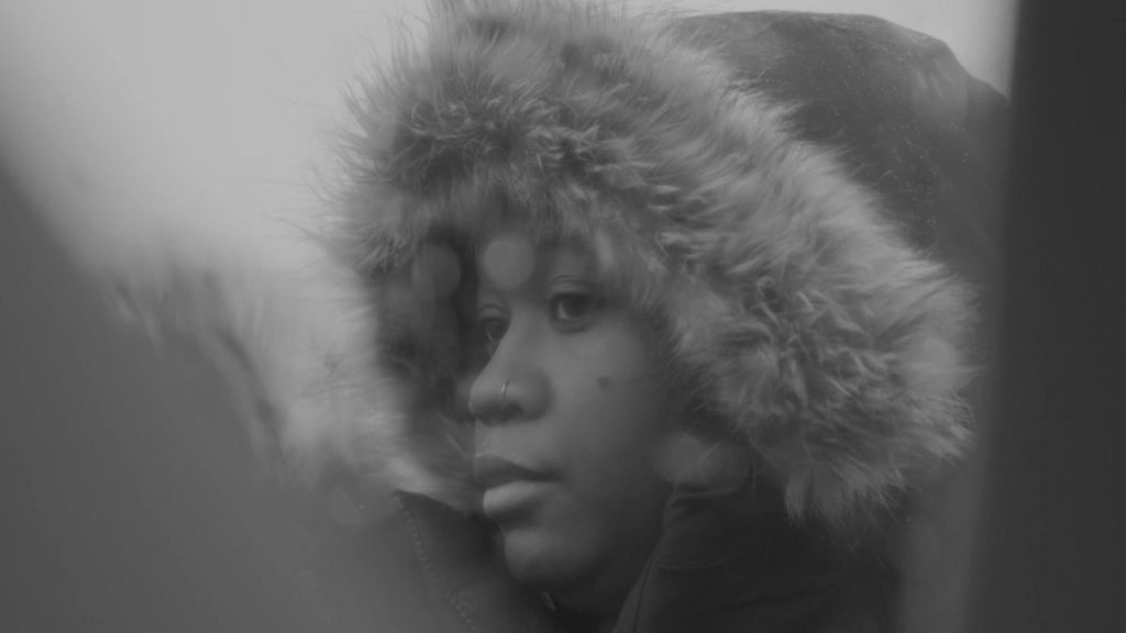 A person wearing a hooded fur-lined coat is gazing thoughtfully. The image is in black and white, with blurred elements in the foreground.
