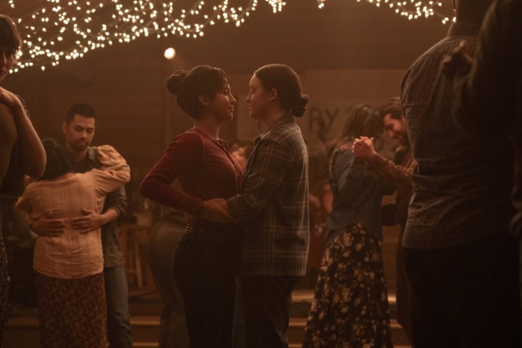 A couple dances closely under string lights at a dimly lit party, surrounded by other dancing pairs.