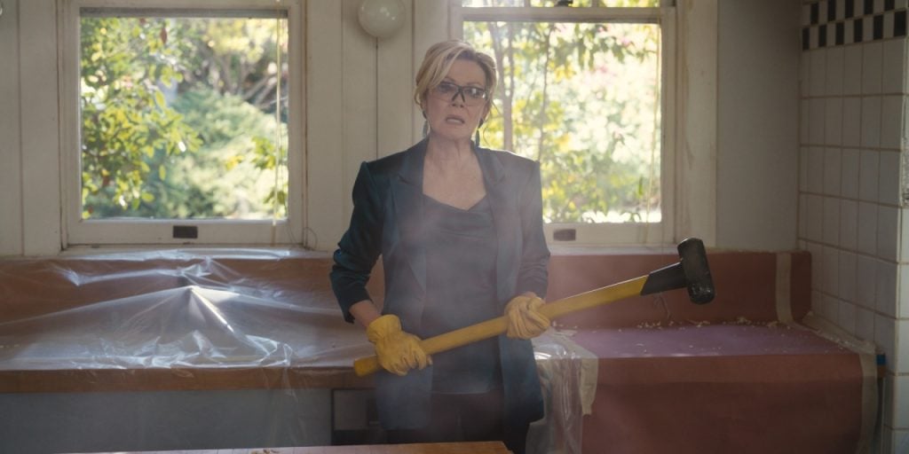 A woman in glasses and gloves stands in a kitchen holding a sledgehammer, with plastic covering surfaces and greenery visible through windows.