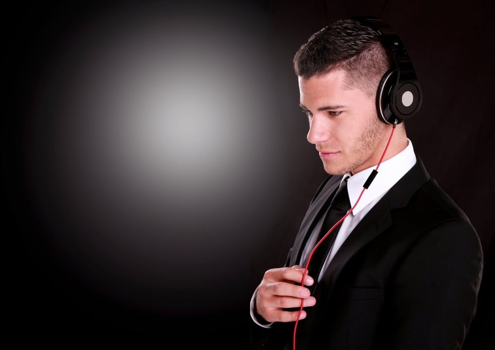 A man in a suit and tie listens to music on headphones with a red cable, standing against a dark background with a bright spot on the left.
