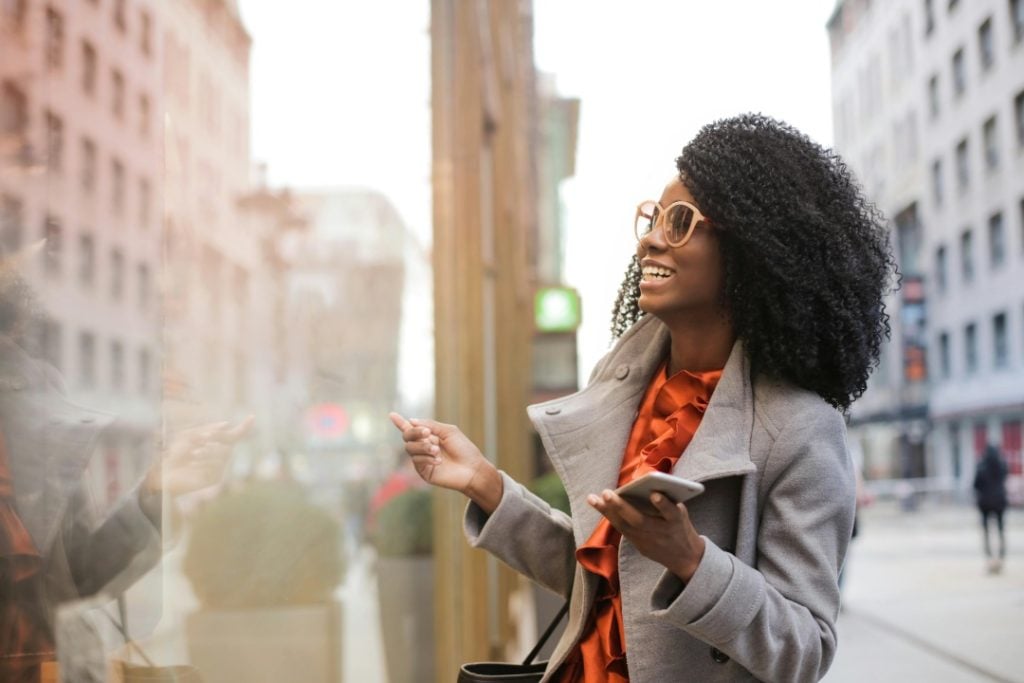 Woman in a gray coat and sunglasses holding a smartphone, smiling and pointing at a storefront window on a city street.