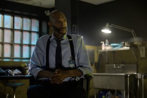 A man in a shirt and tie sits in a dimly lit room with a lamp, sink, and window.