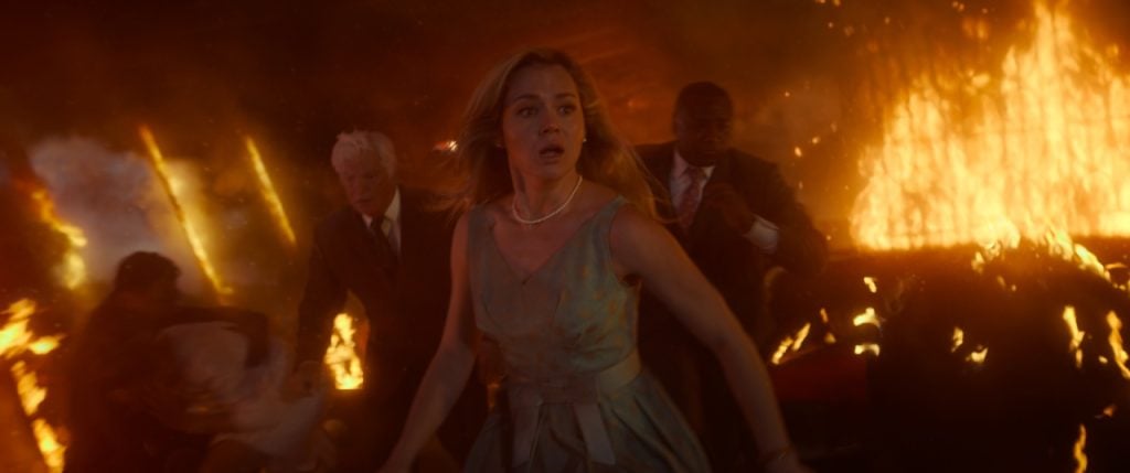 A woman in a dress runs alongside people in suits, fleeing a large fire in an indoor setting.