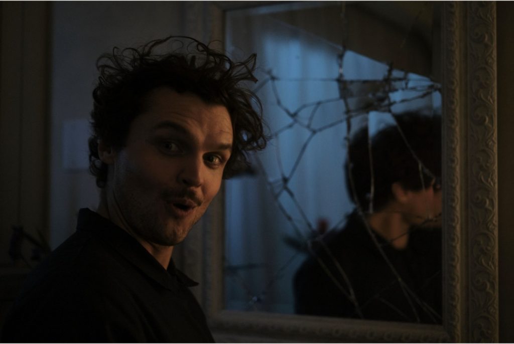 A person with curly hair and a surprised expression stands near a cracked mirror, which reflects their image.