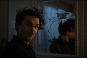 Magnet Releasing To Bring The Samara Weaving & Ray Nicholson Thriller ‘Borderline’ To Blu-Ray This June A person with curly hair and a surprised expression stands near a cracked mirror, which reflects their image.