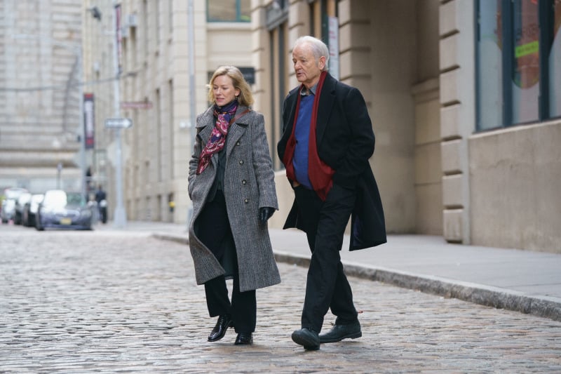 A man and woman walk together on a cobblestone street, both wearing long coats and looking forward. The street is lined with buildings, and a few cars are visible in the background.