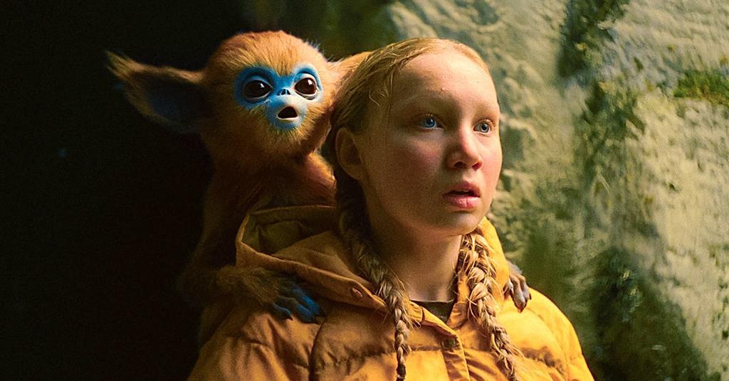 A girl with blonde braids and a yellow jacket stands in a cave with a small, blue-faced, monkey-like creature on her shoulder.
