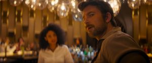 A man in a brown shirt looks toward the camera, with a woman in a white shirt and a blurred background of a bar with glowing lights.