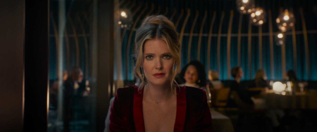 A woman with blonde hair and a serious expression sits at a dimly lit restaurant, wearing a red dress. Other diners are visible in the blurred background.