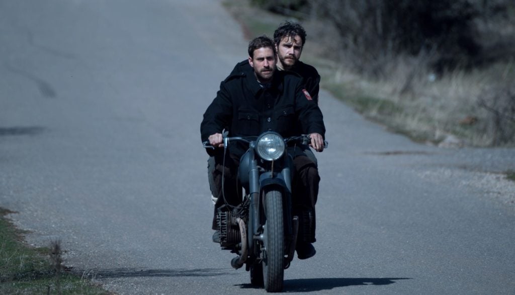 Two men riding a vintage motorcycle down a rural road with grass and trees in the background.