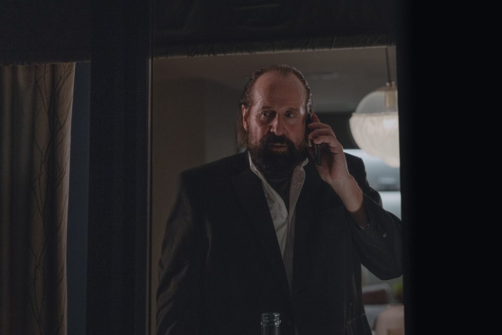 A man with a beard and suit jacket stands indoors, holding a smartphone to his ear and looking slightly concerned.