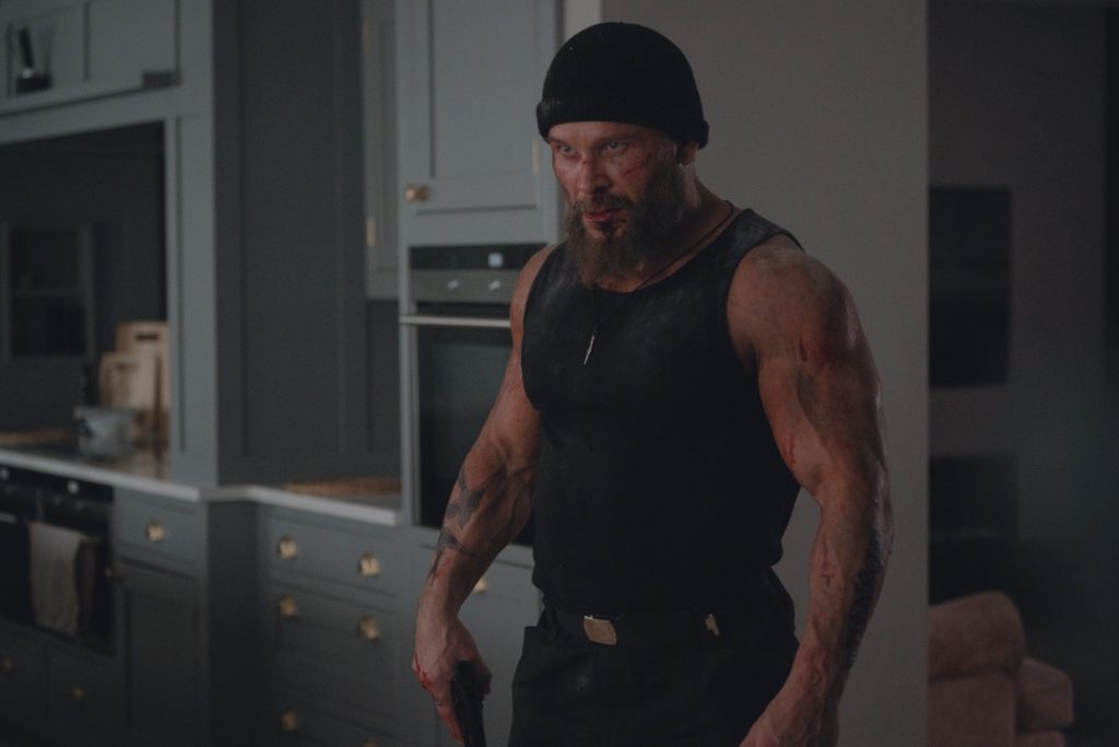A muscular man wearing a black beanie and tank top stands in a kitchen, appearing intense and slightly dirty or injured.
