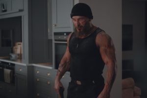 A muscular man wearing a black beanie and tank top stands in a kitchen, appearing intense and slightly dirty or injured.