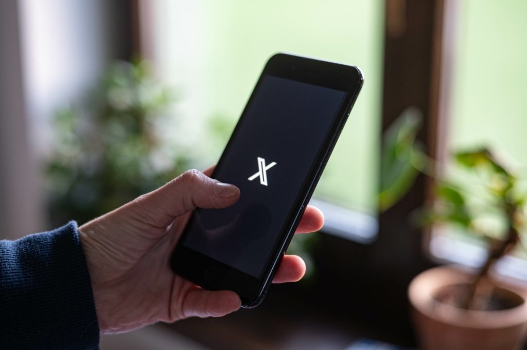 A person holds a smartphone displaying the X logo on the screen, with blurred indoor plants in the background.