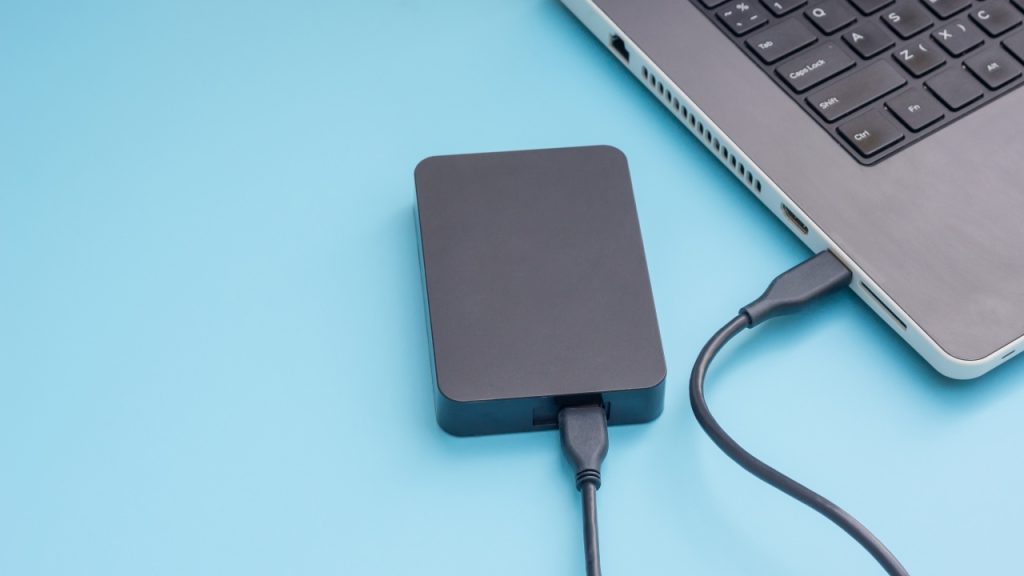 A black external hard drive connected by USB cable to a silver laptop on a blue background.