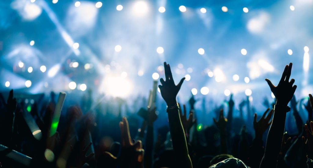 A crowd raises their hands at a concert with bright stage lights and smoke effects illuminating the scene.