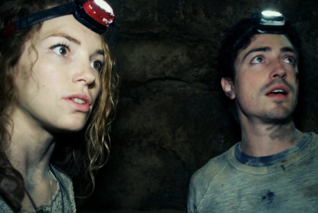 Two people wearing headlamps stand in a dimly lit, rocky environment, looking ahead with alert and concerned expressions.