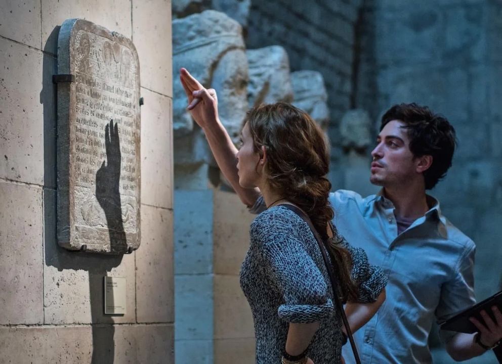 Two people examine a stone tablet mounted on a wall in a museum; one points at the tablet, casting a shadow, while the other holds a tablet device.