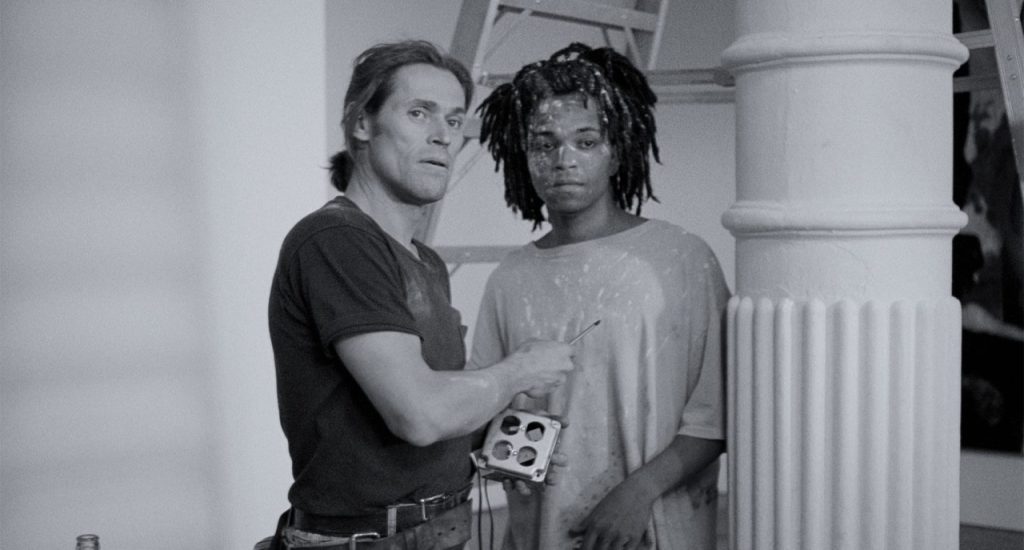 Two artists stand by a pillar; one holds a paintbrush and looks at the camera, while the other, with a paint-splattered face, gazes ahead. A ladder is visible in the background.
