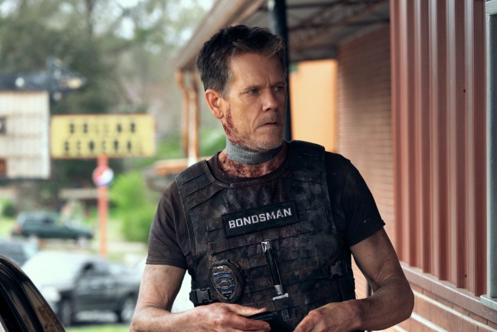 A person wearing a tactical vest labeled "Bondsman" stands outdoors with a vigilant expression, appearing battered and weary.