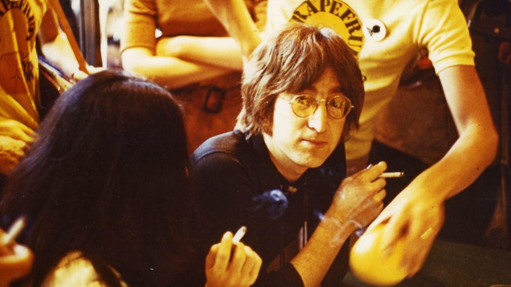 A man with glasses sits at a table holding a cigarette, surrounded by people wearing yellow shirts; another person with long dark hair is in the foreground.