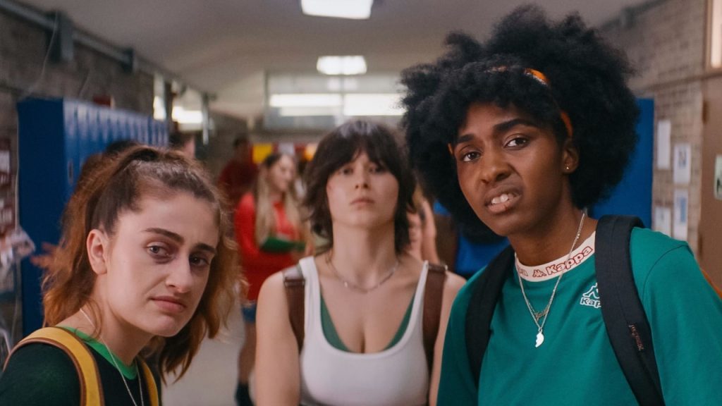Three young women stand in a school hallway, looking directly at the camera with serious and uncertain expressions. Blue lockers and other students are visible in the background.