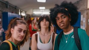 Three young women stand in a school hallway, looking directly at the camera with serious and uncertain expressions. Blue lockers and other students are visible in the background.