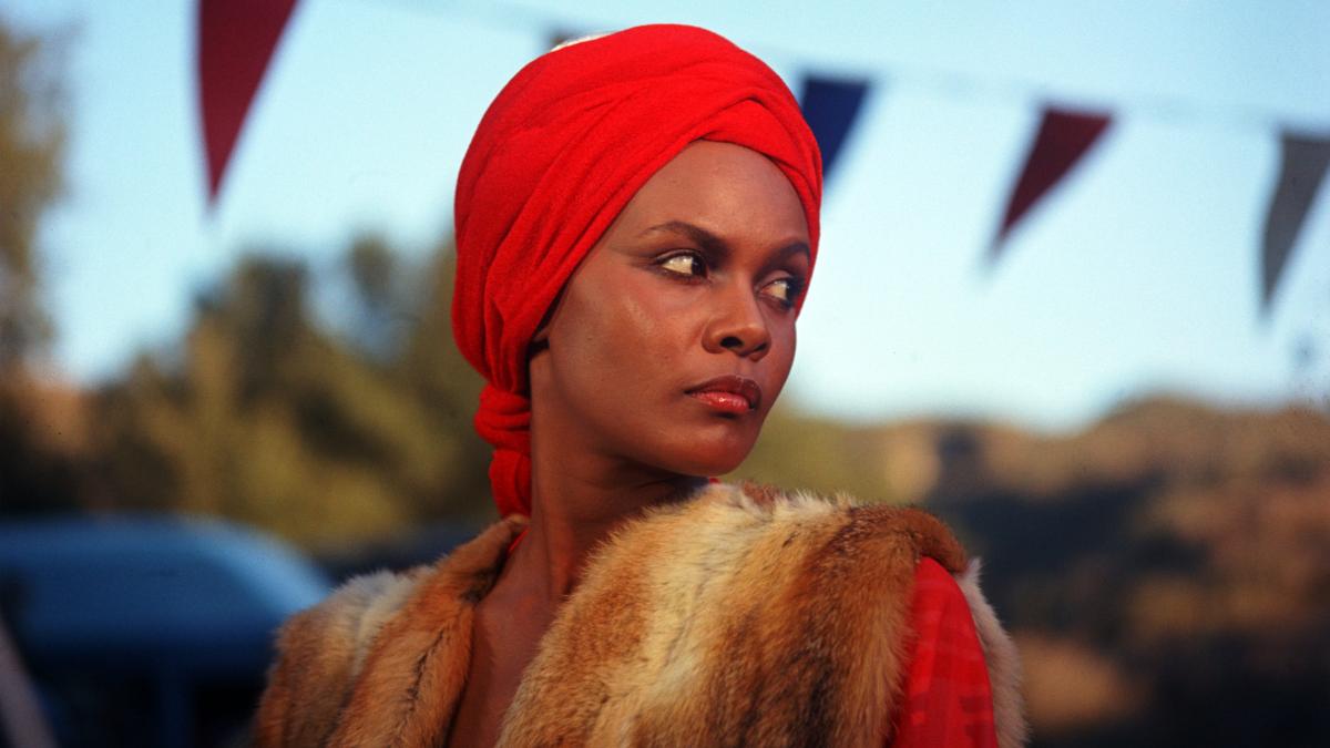 A woman wearing a red headwrap and fur vest looks to the side outdoors with colorful pennant flags in the background.